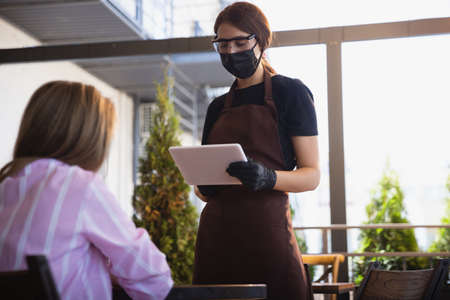 The waitress works in a restaurant in a medical mask, gloves during coronavirus pandemic. Representing new normal of service and safety. Taking orders with tablet contactless. Devices, service.の写真素材