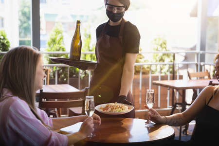 The waitress works in a restaurant in a medical mask, gloves during coronavirus pandemic. Representing new normal of service and safety. Putting the order, taking care of clients, visitors.の写真素材