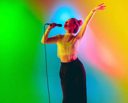 Bright mood. Young female caucasian inspired and expressive musician, singer performing on multicolored background in neon. Concept of music, hobby, festival, art. Joyful artist, colorful portrait.の写真素材