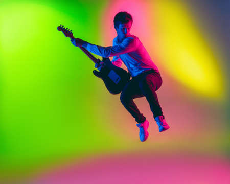 Jump high. Young caucasian inspired and expressive musician, guitarist performing on multicolored background in neon. Concept of music, hobby, festival, art. Joyful artist, colorful, bright portrait.の写真素材