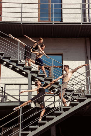 The group of modern ballet dancers performing on the stairs at the city. Fast moving of citylife, grace in daily things around. Graceful and flexible models in sunshine with buildings on background.の写真素材