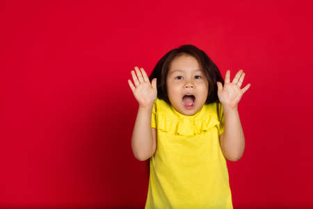 Surprising. Beautiful little girl isolated on red background. Half-lenght portrait of happy child gesturing. Cute asian girl in yellow wear. Concept of facial expression, human emotions, childhood.の写真素材