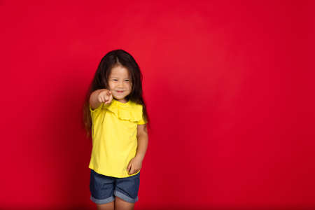 Pointing. Beautiful little girl isolated on red background. Half-lenght portrait of happy child...