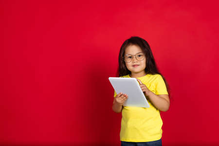Using tablet. Beautiful little girl isolated on red background. Half-lenght portrait of happy child...