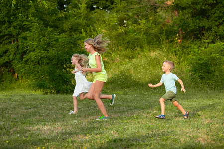 Kids, children running on meadow in summers sunlight. Look happy, cheerful with sincere bright emotions. Cute caucasian boys and girls. Concept of childhood, happiness, movement, family and summer.の写真素材