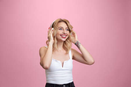 Listening to music with headphones. Caucasian young woman's portrait isolated on pink studio background. Beautiful blonde model. Concept of human emotions, facial expression, sales, ad, youth. Copyspace.の写真素材