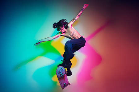 Skateboarder doing a trick isolated on studio background in colorful neon light. Young man shirtless riding and skateboarding in motion. Concept of leisure activity, sport, extreme, hobby and motion.の写真素材