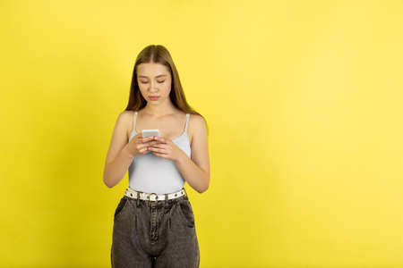 Scrolling phone. Caucasian young girls portrait isolated on yellow studio background. Beautiful female blonde model. Concept of human emotions, facial expression, sales, ad, youth culture. Copyspace.の写真素材