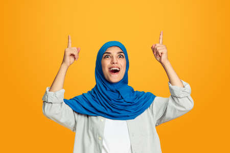 Pointing, smiling. Young muslim woman isolated on yellow studio background. Stylish, trendy and...