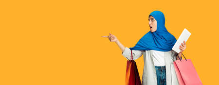 Pointing shocked with tablet. Young muslim woman with shopping packages in casual on yellow studio background. Stylish, trendy. Human emotions, facial expression, sales, ad, shopping concept. Flyer.の写真素材