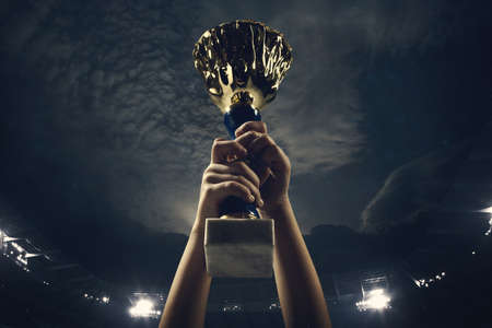 Hard work. Award of victory, male hands tightening the golden cup of winners against cloudy dark sky. Sport, competition, championship, winning, achieving the goal. Prize for success and honor.の写真素材
