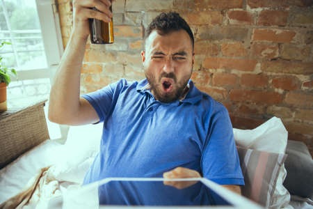 Sport fan, drinks beer. Lazy man living in his bed surrounded with messy. No need to go out to be happy. Using gadgets, watching movie and series, looks emotional. Eating snacks and fast food.の写真素材