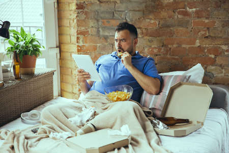 Shocked, biting crisps. Lazy man living in his bed surrounded with messy. No need to go out to be happy. Using gadgets, watching movie and series, looks emotional. Eating snacks and fast food.の写真素材