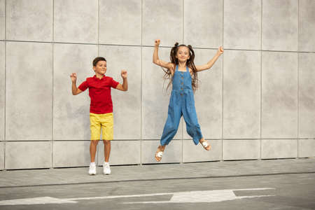 Two smiling kids, boy and girl jumping together in town, city in summer day. Concept of childhood, happiness, sincere emotions, carefree lifestyle. Little caucasian models in bright clothes.の写真素材