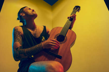 Young musician with guitar performing on yellow background in neon light. Concept of music, hobby, festival, entertainment, emotions. Joyful, inspired guitarist. Colorful portrait of artist.の写真素材