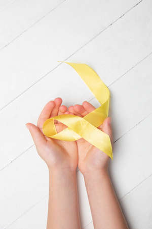 Human hands make yellow ribbon for fighting bone cancer symbol on white wooden background. Concept...