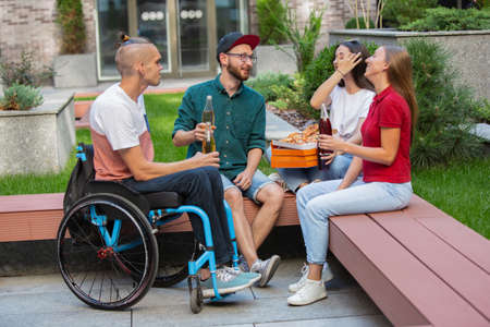 Talking. Group of friends taking a stroll on citys street in summer day. Handicapped man with his friends having fun. Inclusion and diversity concept, normal lifestyle of special groups of society.の写真素材