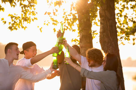 Youth. Group of friends clinking beer bottles during picnic at the beach in sunshine. Lifestyle,...