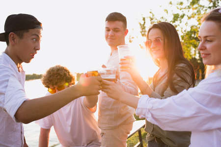 Sunset. Group of friends clinking beer glasses during picnic at the beach in sunshine. Lifestyle, friendship, having fun, weekend and resting concept. Looks cheerful, happy, celebrating, festive.の写真素材