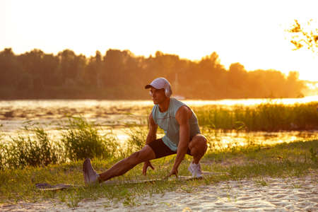 Stretching. A young athletic man working out, training listening to music at riverside outdoors....