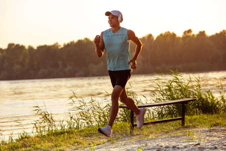 Runner. A young athletic man working out, training listening to music at riverside outdoors. Concept of healthy lifestyle, wellness, sport, activity, weight loss. Inspired of nature, summertime.の写真素材
