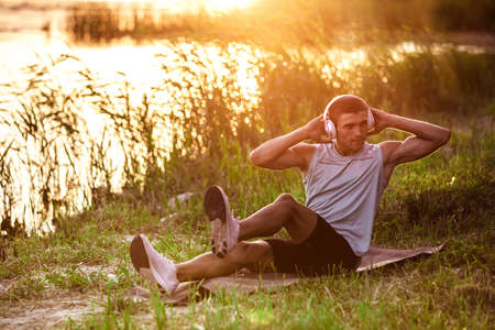 ABS crunches. A young athletic man working out, training listening to music at riverside outdoors. Concept of healthy lifestyle, wellness, sport, activity, weight loss. Inspired of nature, summertime.の写真素材