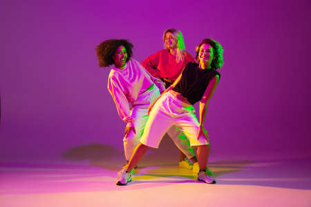 Flexible. Sportive girls dancing hip-hop in stylish clothes on purple-pink background at dance hall in green neon light. Youth culture, movement, style and fashion, action. Fashionable portrait.の写真素材