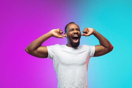Closing ears. African-american young mans portrait on gradient studio background in neon. Beautiful male model in casual style, white shirt. Concept of human emotions, facial expression, sales, ad.の写真素材