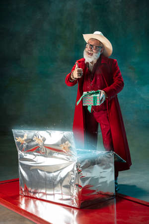 Santas offers. Modern stylish Santa Claus in red fashionable suit and cowboys hat on dark background. Looks like a rockstar. New Year and Christmas eve, celebration, holidays, winters mood, fashion.の写真素材