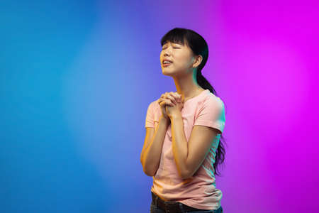 Hopeful praying. Asian young womans portrait on gradient studio background in neon. Beautiful female...