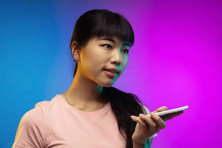 Recording voice. Asian young womans portrait isolated on gradient studio background in neon. Beautiful female model in casual style. Concept of human emotions, facial expression, youth, sales, ad.の写真素材