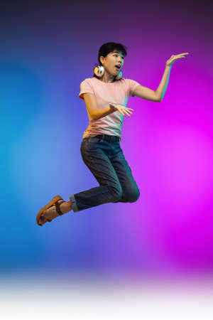 Headphones. Jumping high asian young womans portrait on gradient studio background in neon. Beautiful female model in casual style. Concept of human emotions, facial expression, youth, sales, ad.の写真素材