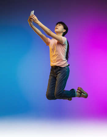Taking selfie. Jumping high asian young womans portrait on gradient studio background in neon. Beautiful female model in casual style. Concept of human emotions, facial expression, youth, sales, ad.の写真素材