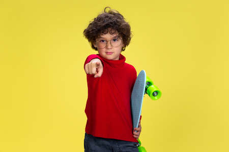 Pointing. Portrait of pretty young curly boy in red wear on yellow studio background. Childhood,...