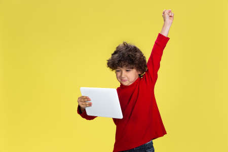 Using tablet. Portrait of pretty young curly boy in red wear on yellow studio background. Childhood,...