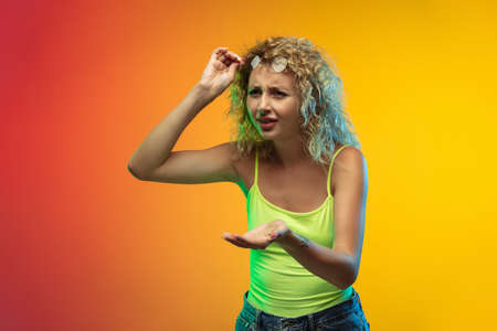Asking. Caucasian young womans portrait isolated on gradient studio background in neon. Beautiful female curly model in casual style. Concept of human emotions, facial expression, youth, sales, ad.の写真素材