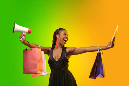Shouting with shopping bags and tablet. Portrait of young woman in neon light on gradient backgound....