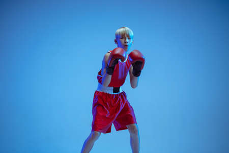 Teenager in sportswear boxing isolated on blue studio background in neon light. Novice male caucasian boxer training hard and working out, kicking. Sport, healthy lifestyle, movement concept.の写真素材
