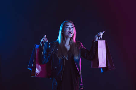 Dancing with shopping bags, cheerful and happy. Portrait of young woman in neon light on dark backgound. The human emotions, black friday, cyber monday, purchases, sales, finance concept.の写真素材