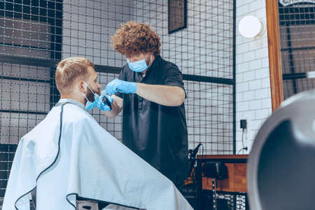 Man getting hair cut at the barbershop wearing mask during coronavirus pandemic. Professional barber wearing gloves. Covid-19, beauty, selfcare, style, healthcare and medicine concept.の写真素材