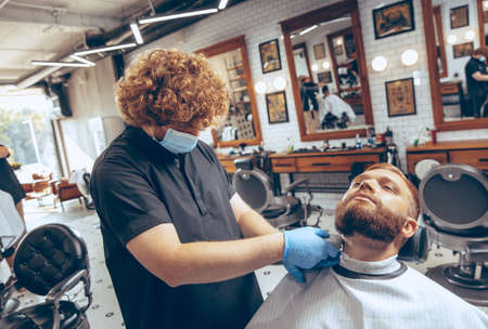 Man getting hair cut at the barbershop wearing mask during coronavirus pandemic. Professional barber wearing gloves. Covid-19, beauty, selfcare, style, healthcare and medicine concept.の写真素材