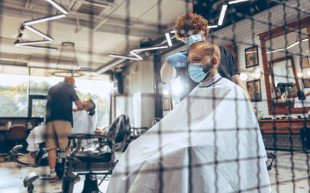 Man getting hair cut at the barbershop wearing mask during coronavirus pandemic. Professional barber wearing gloves. Covid-19, beauty, selfcare, style, healthcare and medicine concept.の写真素材