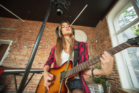Cheerful. Beautiful woman in headphones recording music, singing and playing guitar while sitting in...