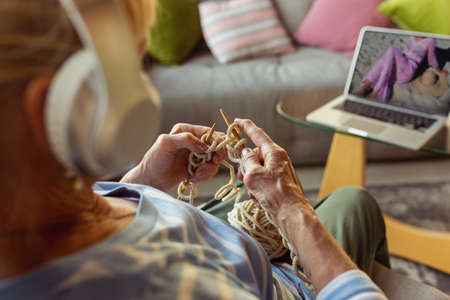 Learning to knit with an online lecturer. Senior woman studying at home, getting online courses, self-development. Caucasian woman using modern devices for education, spending time for new job or hobby.の写真素材