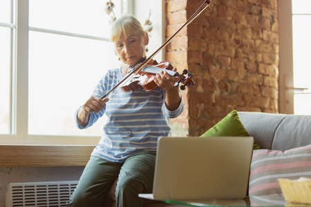 Learning to play violin online. Senior woman studying at home, getting online courses, self-development. Caucasian woman using modern devices for education, spending time for new job or hobby.の写真素材