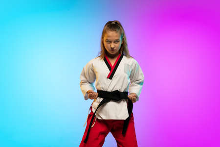 Powerful. Karate, taekwondo girl with black belt isolated on gradient background in neon light. Little caucasian model, sport kid training in motion and action. Sport, movement, childhood concept.の写真素材