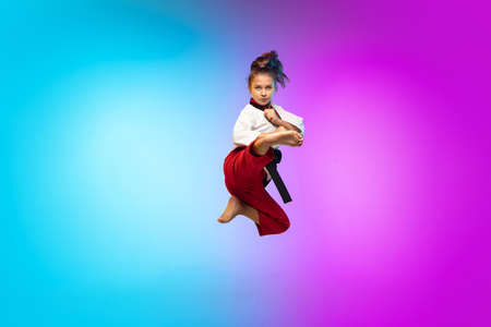 In jump. Karate, taekwondo girl with black belt isolated on gradient background in neon light. Little caucasian model, sport kid training in motion and action. Sport, movement, childhood concept.の写真素材