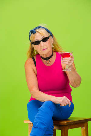 Party time. Senior woman in ultra trendy attire isolated on bright green background. Looks stylish and fashionable, forever young. Caucasian mature woman in sunglasses, bright attire and sneakers.の写真素材