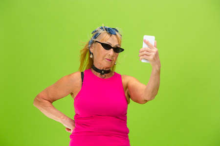 Taking selfie. Senior woman in ultra trendy attire isolated on bright green background. Looks...