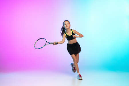 Winner. Little tennis girl in black sportwear isolated on gradient background in neon light. Little...